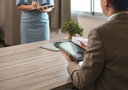 Businessman manager in brown suit using PC tablet while sitting at office desk and secretary in blue dress taking notes while standing in front of his boss. Business work conceptの写真素材
