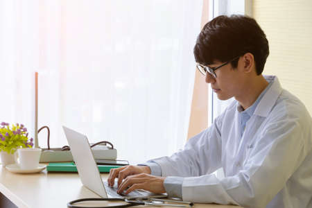 Young doctor physician wearing white uniform and using laptop in hospital office. Male doctor in...