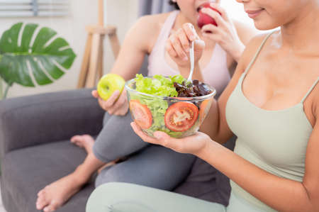 Two young women wearing a sportive outfit and enjoy heathy meal with fruit and vegetable salad on sofa after yoga execise at home with happiness. Healthy lifestyle and exercise conceptの写真素材