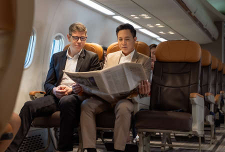 Portrait of two business men reading newspaper in plane enjoying long business flight. Man in brown...