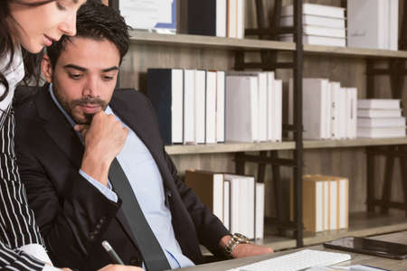Businessman and businesswoman discussing over documents at desk. Businessman sitting in office desk and looking at paperwork while business woman writing and standing next to him.の写真素材