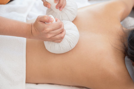 Masseur hands make back massage with Thai herbal compress for young woman on massage tableの写真素材