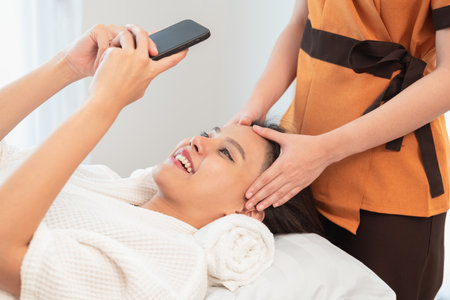 Young smiling woman taking selfie at beauty salon. Beautiful happy girl using smartphone while having spa facial massage.の写真素材