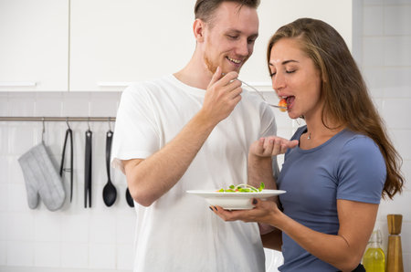 Smiling young couple in love making healthy vegan salad in kitchen,  girl testing it from man hand. Happy family spending time together in weekendの写真素材
