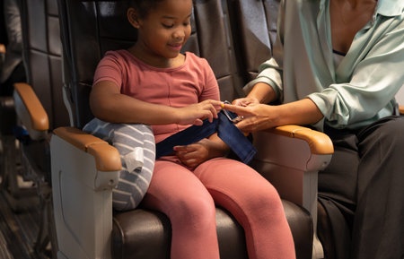 Mother helping her curly daughter to adjust and tight seat belt in airplane cabin for safe flight. Airline transportation and tourism concept.の写真素材