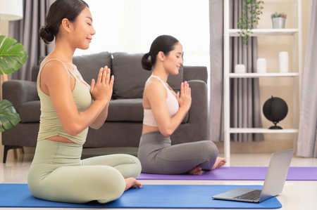 Beautiful two Asian women in sports clothes sitting in lotus position with laptop on blue mat while doing yoga meditating relaxed with closed eyes together at home. Fitness, people and healthy lifestyle and online yoga class conceptの写真素材