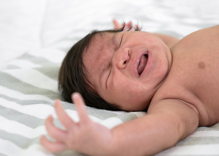 Newborn baby crying on white and grey striped blanket sheet. Infant 1 month screaming on bed. Colic and abdominal pain infant.の写真素材