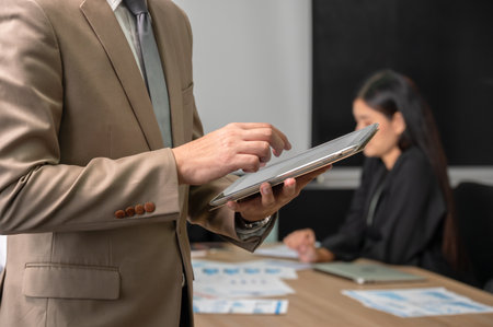 Businessman in brown suit using digital table. Male entrepreneur standing in boardroom with colleagues in backgroundの写真素材
