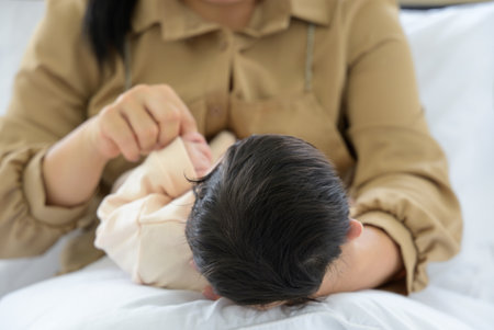 Baby head in mother hand. Mum holding newborn girl lying on white blanket at home. Mom sitting and nursing infant baby on bed. Love, Health Care and mothers day conceptの写真素材