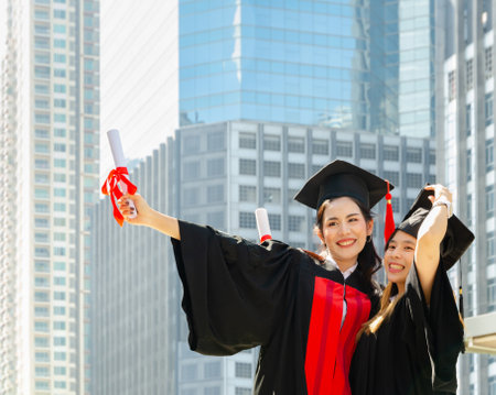 Two cheerful graduated woman wearing mortarboard and academic dress, holding diploma. Smiling women hugging each other on graduation ceremony and standing outdoors with city buildings backgroundの写真素材