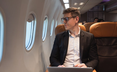 Businessman in formal wear and glasses looking out of aircraft window during flight  while working with laptop computer. Internet connection on board service and business travel conceptの写真素材