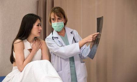 Doctor and patient discussing scan results in hospital. Female doctor checking examining x-ray film of woman patient while sitting on bed at hospital ward.の写真素材