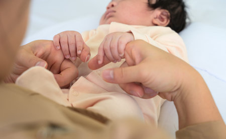 Hands of newborn holding finger of mother hand. Mom sitting on bed and nursing baby girl or boy with love at home. Care, love and protection concept.の写真素材