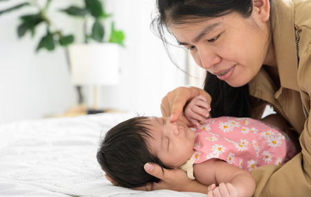 Cute newborn baby girl 1 month old sleeping on mom hand at home. Asian mother on white bed  nursing and gently touching nose of infant baby daughter with love.  Care, love and mothers day concept.の写真素材