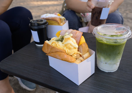 Breakfast toast sandwich with bacon, egg and cheese in take away box and cold drinks on wooden table at the parkの写真素材