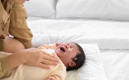 Baby newborn girl crying with mother comforting in bed at home. Mom nursing newborn baby girl and gently touching on chest with love. Healthcare and mother's dayの写真素材
