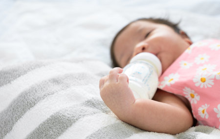 Hand of newborn baby 1 month old holding plastic milk bottle. Infant girl lying alone in bed and...