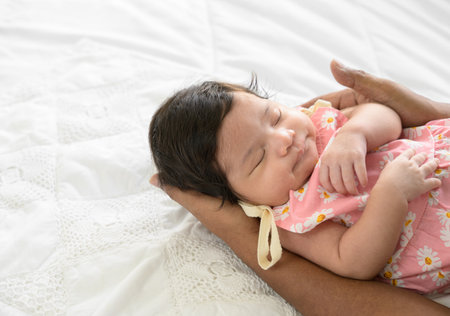 Charming newborn baby girl 1 month old sleeping on dad hand at home. Father nursing infant on white bed and gently touching her  with love. Care, love and fathers day concept.の写真素材