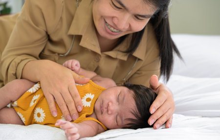 Asian smiling mom motherhood lying down nursing newborn baby girl and gently touching on chest and head with love. Infant sleep comfortable on white bed. Care, love and protection concept.の写真素材