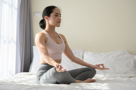 Asian female in sports clothes sitting in lotus position on bed. Woman doing yoga exercise meditating relaxed with closed eyes at home. Morning workout in bedroom. Healthy, relaxation and sport lifestyle.の写真素材