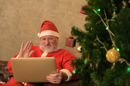 Happy senior caucasian Santa Claus using laptop and waving hand on video calling to child and greeting on Merry Christmas and Happy New Year in virtual video online chat while sitting on sofa near decorated Christmas tree in living room at home with presents on xmas eve.の写真素材