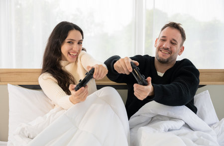 Smiling young couple in sweater playing video games with controller in bed. Man and woman having fun and enjoying together at home. Gaming and technology conceptの写真素材