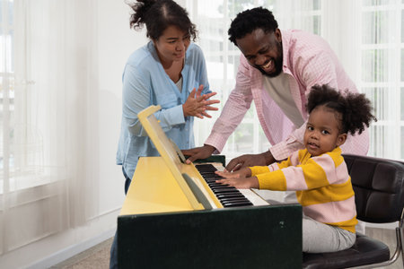 Happy family playing piano at home. African father teaching little curly daughter playing piano in...