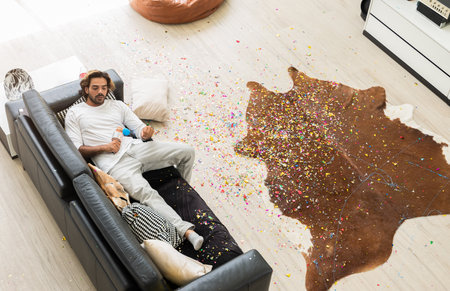 Man lying on black leather couch and holding microphone in disheveled room with colorful confetti on wooden floor after Christmas celebration party at home.の写真素材