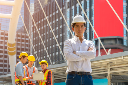 Portrait of Asian male project engineer in white helmet standing with crossed arms in front of modern building with three men working with laptop on backgroundの写真素材
