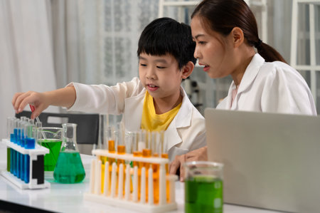 Boy in white lab coat and yellow shirt conducting science experiment and handling test tubes while sitting with Asian woman teacher or mentor. Colorful liquids in beakers and flasks on table in laboratory room. Children education concept with experiment, fun and enjoy class.の写真素材