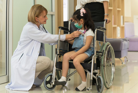 Female doctor kneeling and examining a little girl with a stethoscope at hospital. Asian kid with medical facial mask in a wheelchair. Medicine and healthcare services concept.の写真素材