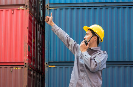 Japanese foreman in yellow hard hat and grey uniform gives thumbs up gesture while speaking on...