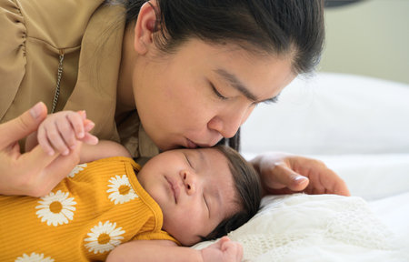 Asian young mom kissing her cute newborn little baby girl sleeping in white bed at home. Healthcare, love, protection and mother's day conceptの写真素材