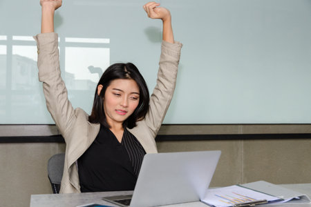 Relaxed Asian businesswoman in tan blazer stretching arms overhead at office desk while looking down at laptop with satisfied or tired expressionの写真素材
