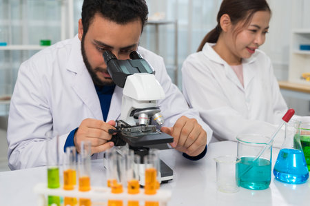 Male scientist with beard examining sample through microscope while female colleague working nearby in science laboratory with colorful chemical solutions in test tubes and beakersの写真素材