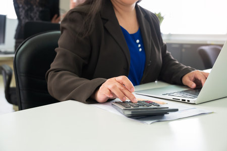 Businesswoman in suit using calculator while working on laptop at office desk. Financial analysis...