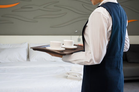 Hotel woman staff member in uniform holding tray with two white cups and saucers while providing room service in  neatly arranged modern guest room with freshly made bed in background.の写真素材