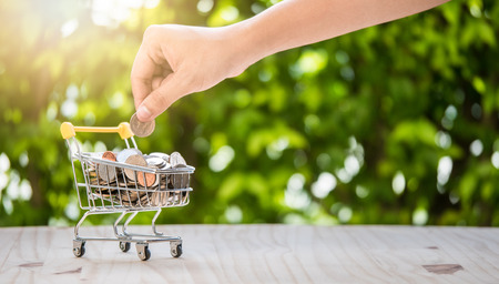 Hand holding coin put in shopping cart on wood table with bokeh background in finacial or business conceptの写真素材