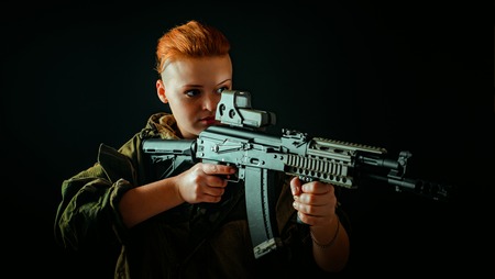 Portrait of young woman with red hair, young girl takes aim at the sight in military uniformの写真素材