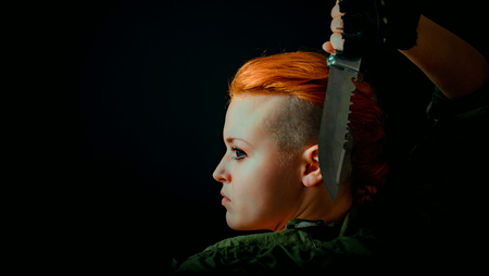 Young woman with red hair stand back in military uniform, keep a knife in hand. Horizontal photoの写真素材