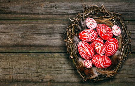 Easter red eggs with folk white pattern inside bird nest on right side of rustic wood board. Top view.の写真素材