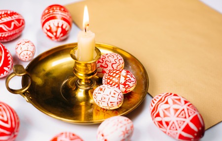 Red easter eggs with folk white pattern around candlestick with burning candle on paper on white background. Ukrainian traditional eggs pisanka and krashanka.の写真素材