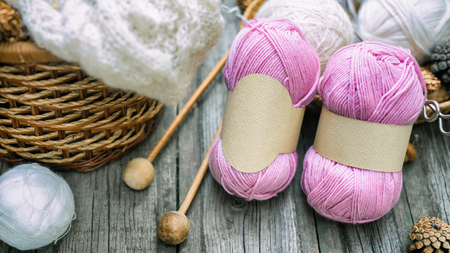 Two pink balls of wool threads with empty labels lying on vintage wood desk with knitting needles and white ball roll to basketの写真素材