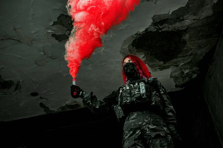 Airsoft red-hair woman in uniform with red smoke in hand. Close up soldier in broken building. Horizontal photoの写真素材