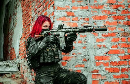 Airsoft red-hair woman in uniform with machine gun beside brick wall. Soldier aims at the sight on the ruins. Side viewの写真素材