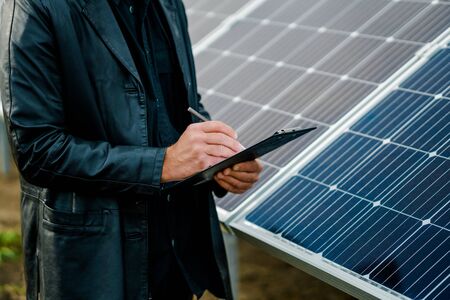 Closeup man write notes on clipboard beside solar battery.の写真素材