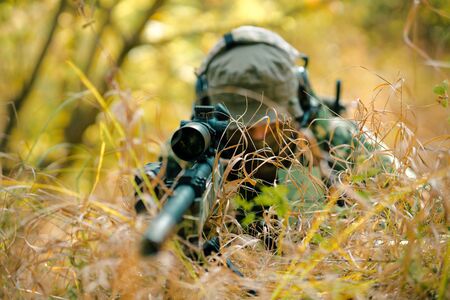 Closeup man in uniform with sniper rifle, front view. Airsoft soldier lay in long grass and check wind correction in note.の写真素材