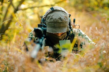 Closeup man in uniform with sniper rifle, front view. Airsoft soldier lay in long grass and check wind correction in note.の写真素材