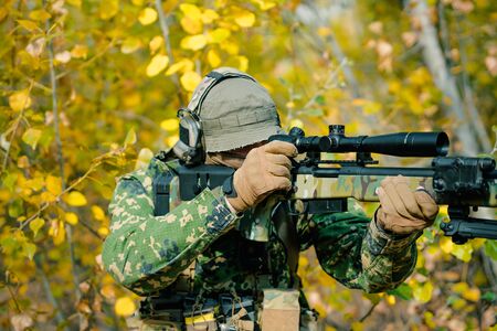 Airsoft man in uniform with sniper rifle, make correction in optical sight on yellow forest backdrop. Soldier aims at the sight.の写真素材