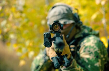 Closeup airsoft man in uniform, stand with sniper rifle on yellow forest backdrop. Soldier aims at the sight. Front view.の写真素材